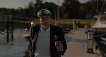 Movie still from “Confess, Fletch” (2022), directed by Greg Mottola – An older man wearing a sailor's hat and holding a drink; Medium shot, Over the shoulder angle