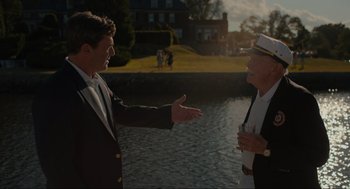 Movie still from “Confess, Fletch” (2022), directed by Greg Mottola – Two men in suits are talking next to a body of water; Medium shot, Over the shoulder angle