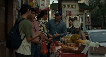 Movie still from “Confess, Fletch” (2022), directed by Greg Mottola – A group of people standing around a fruit stand; Medium shot, Over the shoulder angle