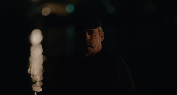 Movie still from “Confess, Fletch” (2022), directed by Greg Mottola – A man in a black shirt and a black hat is in the dark; Medium shot, Low angle