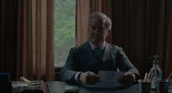 Movie still from “Confess, Fletch” (2022), directed by Greg Mottola – An older man sitting at a table holding papers; Close Up shot, Over the shoulder angle