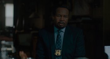 Movie still from “Confess, Fletch” (2022), directed by Greg Mottola – A man in a suit and tie wearing a police badge; Close Up shot, Over the shoulder angle