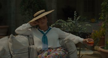Movie still from “Confess, Fletch” (2022), directed by Greg Mottola – A woman sitting in a chair wearing a straw hat and holding her hand up to her head; Medium shot, Over the shoulder angle
