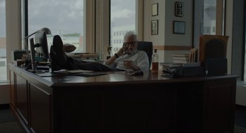Movie still from “Confess, Fletch” (2022), directed by Greg Mottola – An older man sitting at a desk in front of a window; Wide shot, Low angle