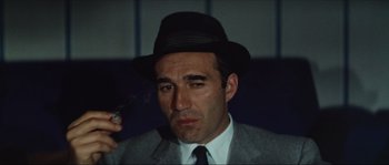 Movie still from “Contempt” (1963), directed by Jean-Luc Godard – A man wearing a suit and a hat smoking a cigarette; Close Up shot, Low angle