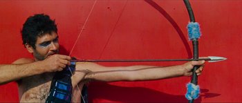 Movie still from “Contempt” (1963), directed by Jean-Luc Godard – A person holding a bow and arrow on the side of a red wall; Medium shot, Low angle