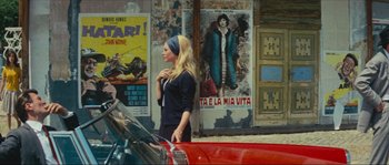 Movie still from “Contempt” (1963), directed by Jean-Luc Godard – A woman walking down a street next to a red car; Medium shot, Over the shoulder angle