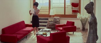 Movie still from “Contempt” (1963), directed by Jean-Luc Godard – A woman standing in a living room with red chairs; Wide shot, High angle