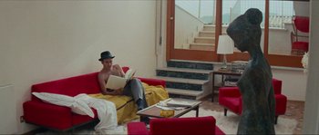 Movie still from “Contempt” (1963), directed by Jean-Luc Godard – A man sitting on top of a red couch in a living room; Wide shot, High angle