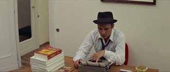 Movie still from “Contempt” (1963), directed by Jean-Luc Godard – A man wearing a hat and a tie is typing on a typewriter; Medium shot, High angle