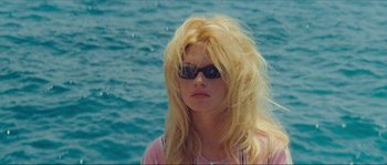 Movie still from “Contempt” (1963), directed by Jean-Luc Godard – A woman with blonde hair and sunglasses standing in front of the ocean; Close Up shot, High angle