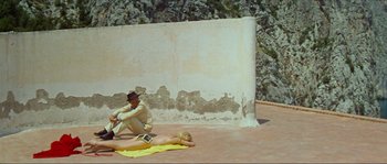 Movie still from “Contempt” (1963), directed by Jean-Luc Godard – A man and a woman laying on the ground in front of a stone wall; Wide shot, High angle