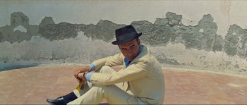 Movie still from “Contempt” (1963), directed by Jean-Luc Godard – A man sitting on the ground wearing a suit and a hat; Medium shot, High angle