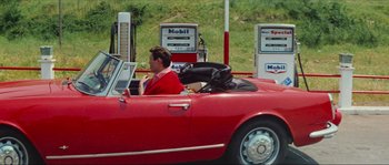 Movie still from “Contempt” (1963), directed by Jean-Luc Godard – A man driving a red car with a suit on; Wide shot, High angle