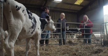 Movie still from “Cow” (2021), directed by Andrea Arnold – Two women are standing in a barn with a cow; Medium shot, Over the shoulder angle