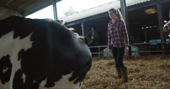Movie still from “Cow” (2021), directed by Andrea Arnold – A woman standing next to a black and white cow in a barn; Medium shot, Over the shoulder angle