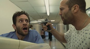 Movie still from “Crank” (2006), directed by Mark Neveldine – A man holding a gun pointed at another man in a hospital hallway; Close Up shot, Over the shoulder angle