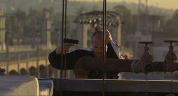 Movie still from “Crank” (2006), directed by Mark Neveldine – A man holding a handgun while standing next to a building; Medium shot, Low angle