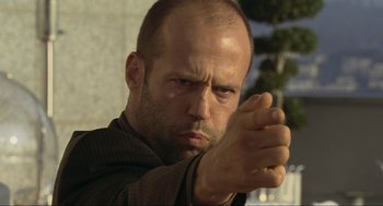 Movie still from “Crank” (2006), directed by Mark Neveldine – A man with a shaved head and a bald head is holding his fist up; Close Up shot, Low angle