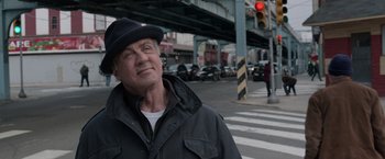 Movie still from “Creed” (2015), directed by Ryan Coogler – A man wearing a black hat and a black jacket crosses the street; Close Up shot, Low angle
