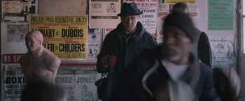 Movie still from “Creed” (2015), directed by Ryan Coogler – A man in a black jacket and a black hat is holding a cup; Medium shot, Over the shoulder angle