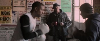 Movie still from “Creed” (2015), directed by Ryan Coogler – A man in a black hat is holding a cup; Medium shot, Over the shoulder angle