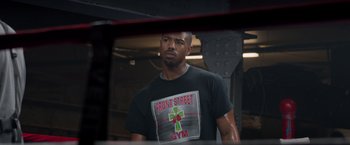 Movie still from “Creed” (2015), directed by Ryan Coogler – A man in a black t - shirt is standing in front of a mirror; Medium shot, Over the shoulder angle