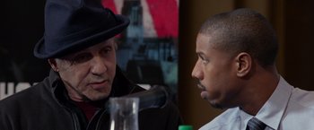 Movie still from “Creed” (2015), directed by Ryan Coogler – A man wearing a black hat and talking to another man in front of a microphone; Close Up shot, Over the shoulder angle