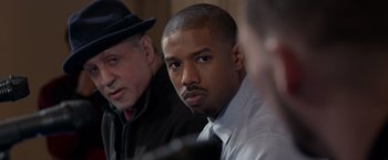 Movie still from “Creed” (2015), directed by Ryan Coogler – A man in a suit and tie sitting next to another man in a suit and tie; Close Up shot, Over the shoulder angle