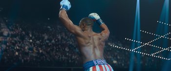 Movie still from “Creed II” (2018), directed by Steven Caple Jr. – A man with his hands raised in a boxing ring; Medium shot, Over the shoulder angle
