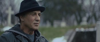 Movie still from “Creed II” (2018), directed by Steven Caple Jr. – An older man wearing a black hat and a gray jacket; Close Up shot, Over the shoulder angle