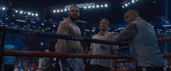 Movie still from “Creed II” (2018), directed by Steven Caple Jr. – A man standing in a boxing ring next to a referee; Medium shot, Over the shoulder angle