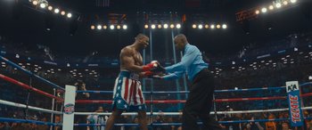 Movie still from “Creed II” (2018), directed by Steven Caple Jr. – Two men in a boxing ring one of them is wearing a blue shirt and the other is wearing a red and white striped shirt; Wide shot, Over the shoulder angle