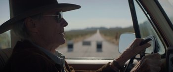 Movie still from “Cry Macho” (2021), directed by Clint Eastwood – An older man driving a car on a road; Close Up shot, Over the shoulder angle
