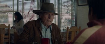 Movie still from “Cry Macho” (2021), directed by Clint Eastwood – An old man wearing a hat sitting at a table with a red cup; Close Up shot, Over the shoulder angle