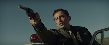 Movie still from “Cry Macho” (2021), directed by Clint Eastwood – A man holding a gun in his hand while standing next to a car; Medium shot, Low angle