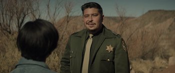 Movie still from “Cry Macho” (2021), directed by Clint Eastwood – A man in a sheriff's uniform standing in front of some trees; Medium shot, Over the shoulder angle