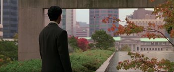 Movie still from “Crying Freeman” (1995), directed by Christophe Gans – A man in a suit looking out a window at a city; Medium shot, Over the shoulder angle