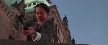 Movie still from “Crying Freeman” (1995), directed by Christophe Gans – A man in a suit and tie standing in front of a building; Close Up shot, Low angle