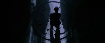 Movie still from “Crying Freeman” (1995), directed by Christophe Gans – A man standing in a dark room with a clock on the wall behind him; Wide shot, Overhead angle