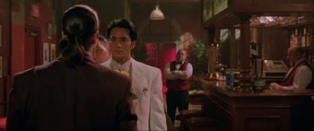 Movie still from “Crying Freeman” (1995), directed by Christophe Gans – A man in a white suit standing next to a bar; Medium shot, Over the shoulder angle