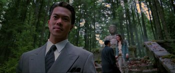 Movie still from “Crying Freeman” (1995), directed by Christophe Gans – A man wearing a suit and tie standing in front of a statue; Close Up shot, Low angle
