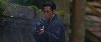 Movie still from “Crying Freeman” (1995), directed by Christophe Gans – A man in a black robe holding a black object in his hands; Close Up shot, Low angle