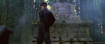 Movie still from “Crying Freeman” (1995), directed by Christophe Gans – A man standing in front of a stone statue; Medium shot, Low angle