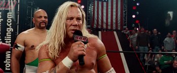 Movie still from “The Wrestler” (2008), directed by Darren Aronofsky – A man with long blond hair is holding a microphone; Close Up shot, Low angle