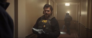 Movie still from “To Catch a Killer” (2023), directed by Damián Szifron – A man wearing a fbi vest holding papers in a hallway; Medium shot, Over the shoulder angle