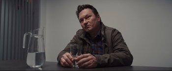 Movie still from “To Catch a Killer” (2023), directed by Damián Szifron – A man sitting at a table holding a glass of water; Medium shot, Over the shoulder angle