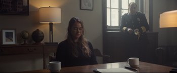 Movie still from “To Catch a Killer” (2023), directed by Damián Szifron – A woman sitting at a wooden table in front of a lamp; Medium shot, Over the shoulder angle