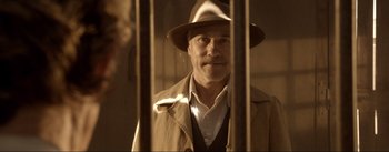 Movie still from “Dead for a Dollar” (2022), directed by Walter Hill – A man wearing a hat and a trench coat standing behind bars; Close Up shot, Low angle