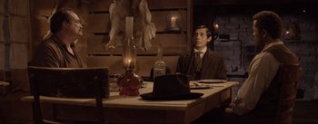 Movie still from “Dead for a Dollar” (2022), directed by Walter Hill – A man sitting at a dinner table with a hat on his head; Medium shot, Over the shoulder angle